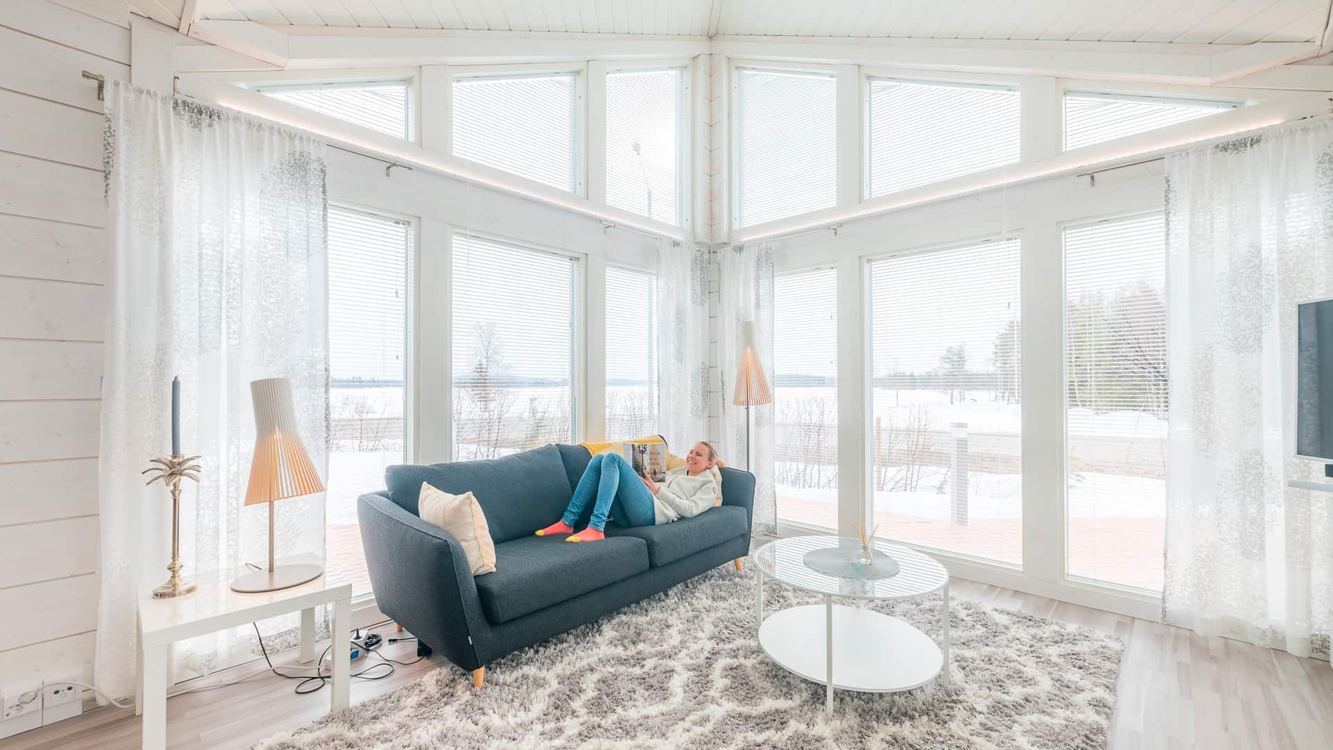 The large windows in the living room of the small log home provide plenty of light. There is a sofa in front of the window, with a person sitting on it reading a book.