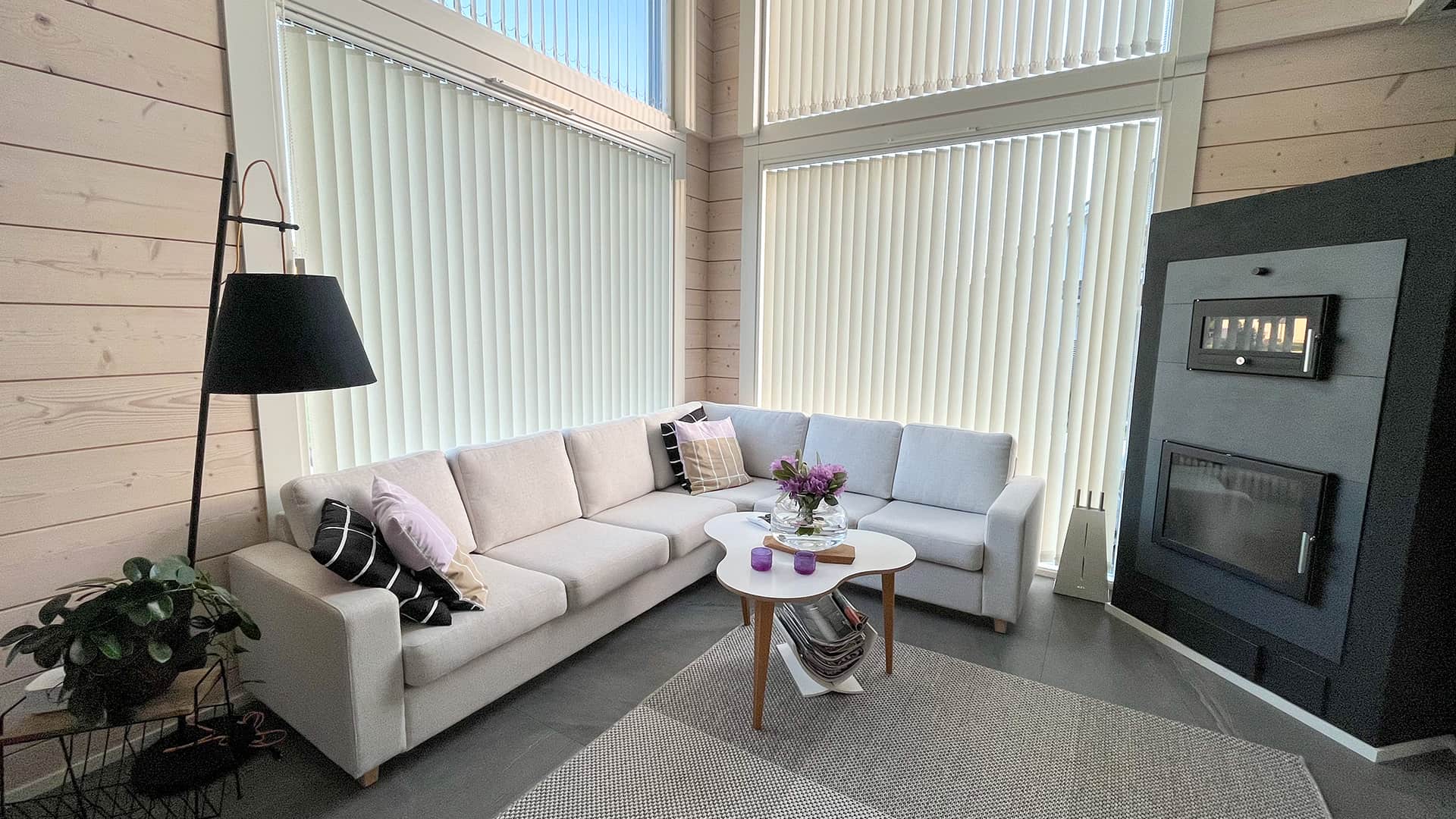 The atmosphere of the open living area in Villa Hiisi There is a lightly coloured corner sofa in front of the large windows in the corner of the living room. There is a dark grey baking oven fireplace in the opposite corner.