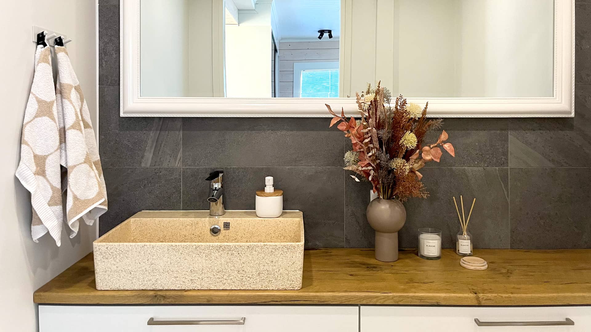 The toilet has grey tile walls behind the sink. There is enough room for toiletries on the top. There is a large mirror with a white frame on the wall.