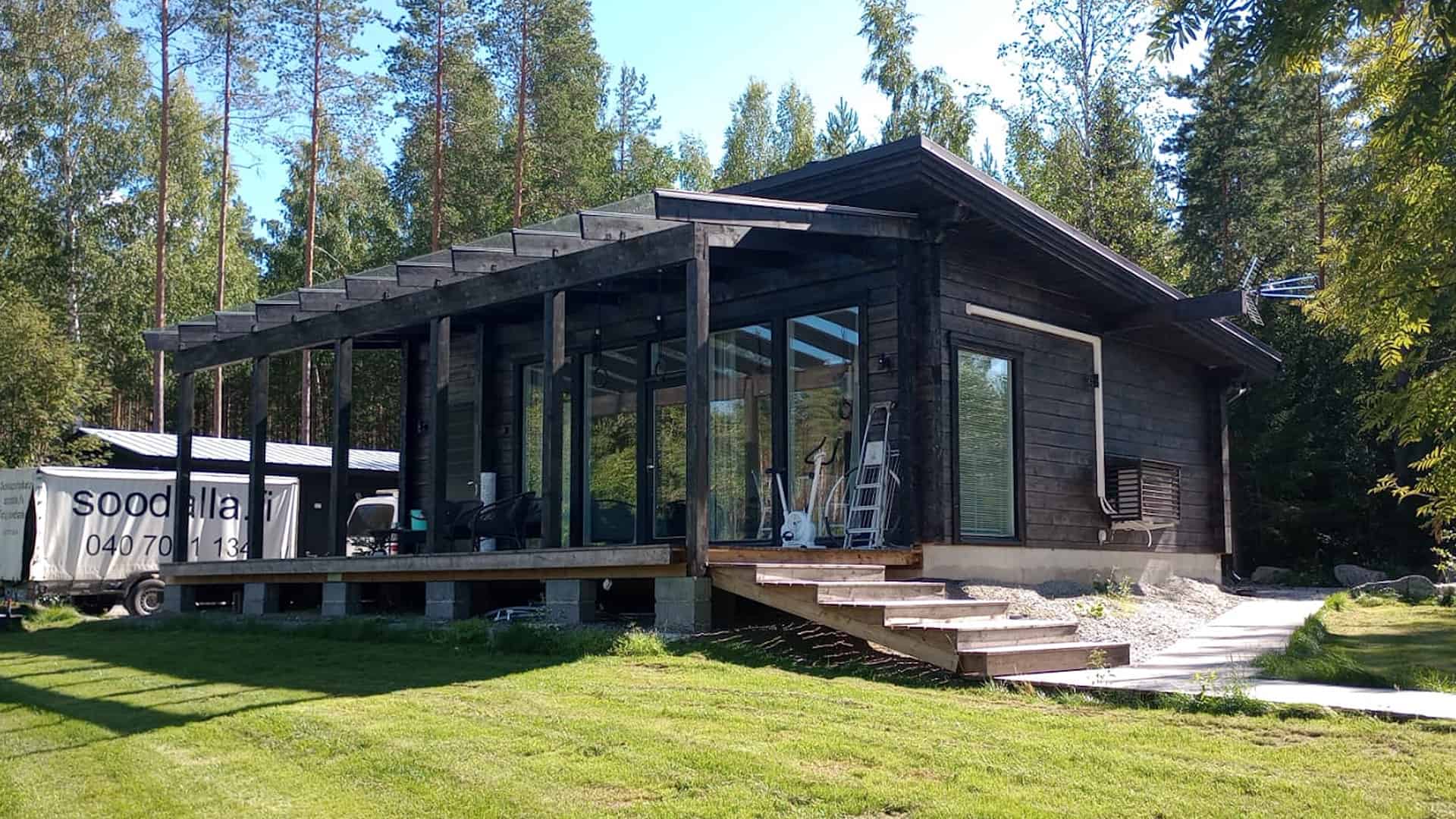 Tervetuloa hirsihuvilanäyttelyyn! Ulkokuva pulpettikattoisesta, mustaksi maalatusta hirsihuvilasta. Edessä on pergolan alla iso terassi.
