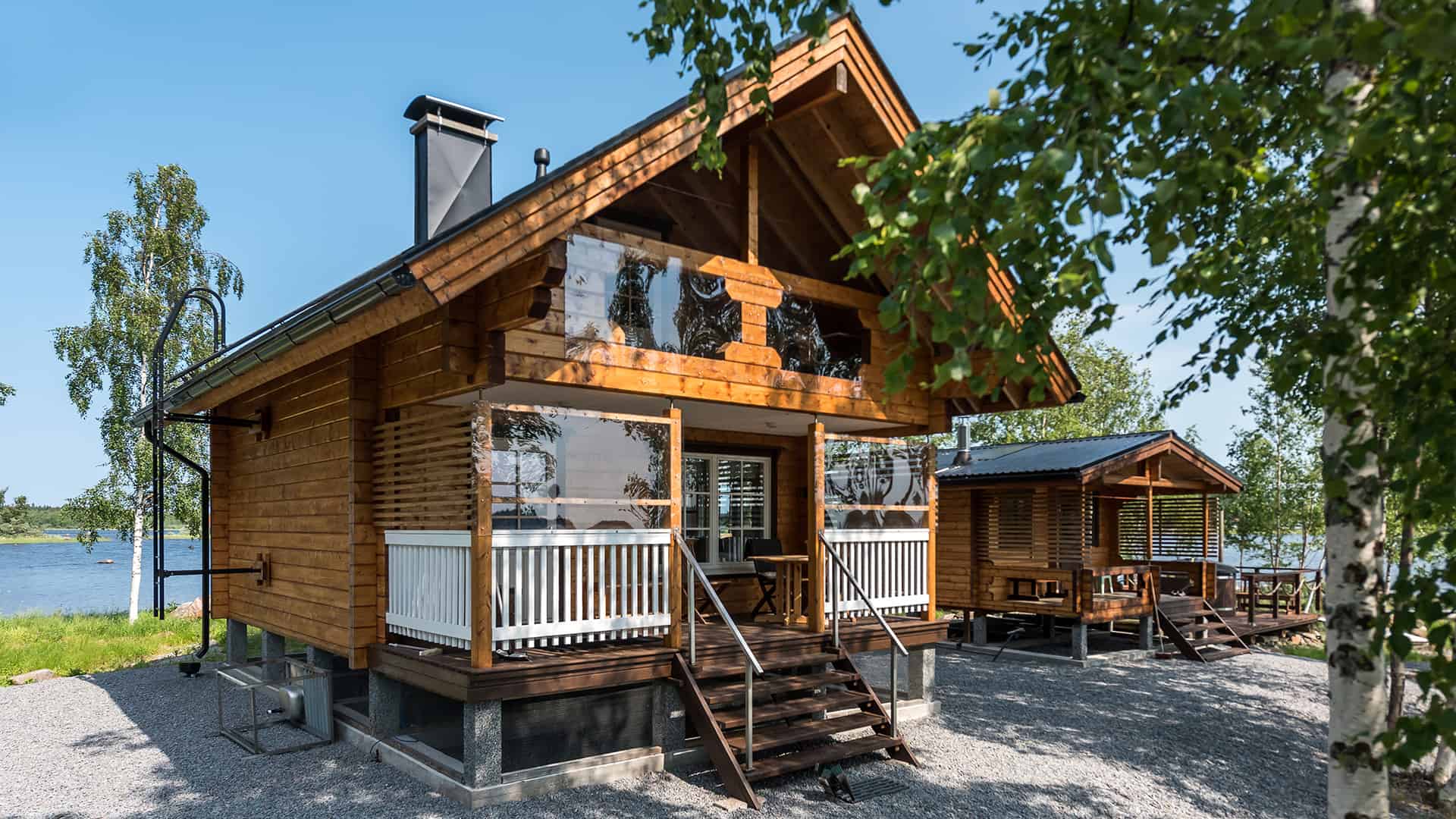 Outside view shows the traditional, ridged roof log cabin by the lake. There is a balcony under the roof and a terrace by the entrance.