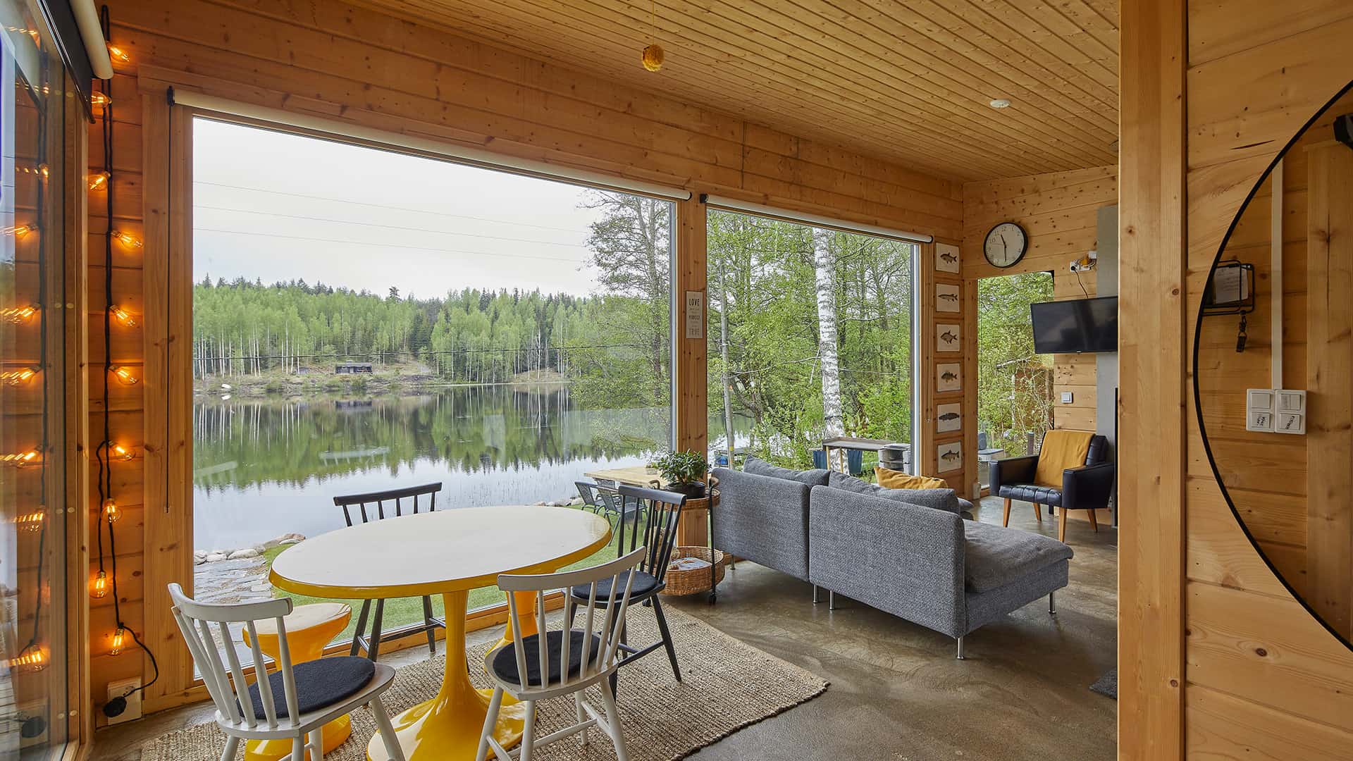 The lounge of the modern log cabin has large windows that cover almost the entire wall. They open up to a lakeside view. The lounge includes a round dinner table for four and a sofa and two armchairs.