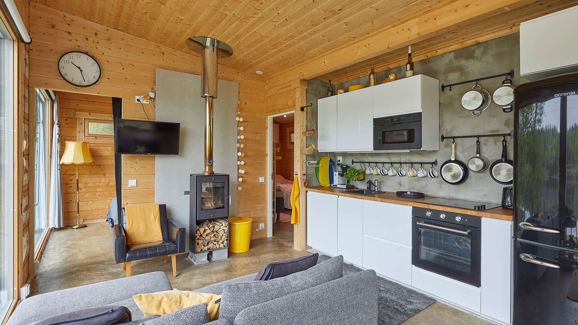 The compact lounge in the log cabin. The white kitchen by Ikea is on the right wall. There is a small stove in the middle of the picture. A slanted ceiling creates a sense of space. 