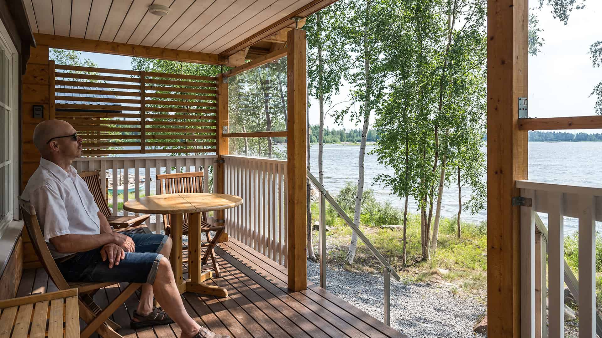 Heikki istuu hirsimökin terassilla katsellen järvimaisemaa. 